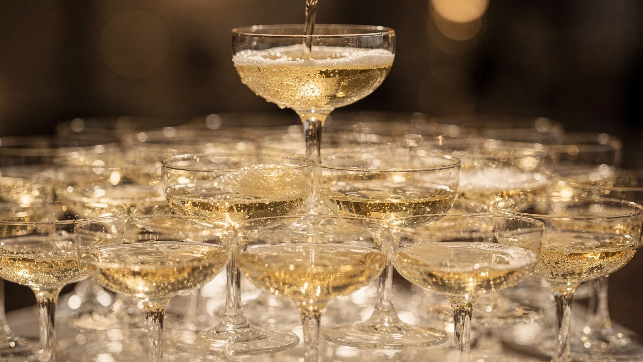 A close-up of a champagne glass pyramid with crystal-clear coupes aligned in perfect rows, soft golden reflections in the glass, and a single stream of champagne being poured into the top glass.
